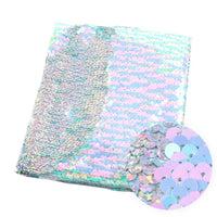 Un morceau plié de Longan Craft France Tissu pailleté bicolore réversible au mètre présente des paillettes rose pastel, bleues et argentées. Un empiècement met en valeur les paillettes rondes et superposées de ce tissu réversible scintillant.