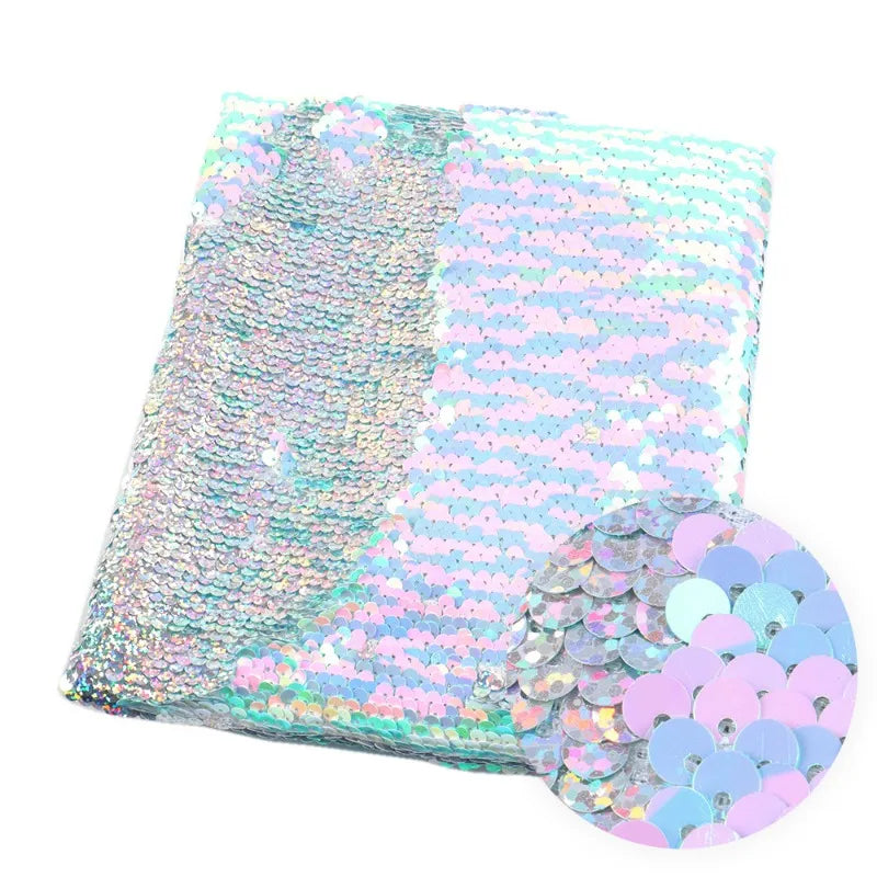 Un morceau plié de Longan Craft France Tissu pailleté bicolore réversible au mètre présente des paillettes rose pastel, bleues et argentées. Un empiècement met en valeur les paillettes rondes et superposées de ce tissu réversible scintillant.