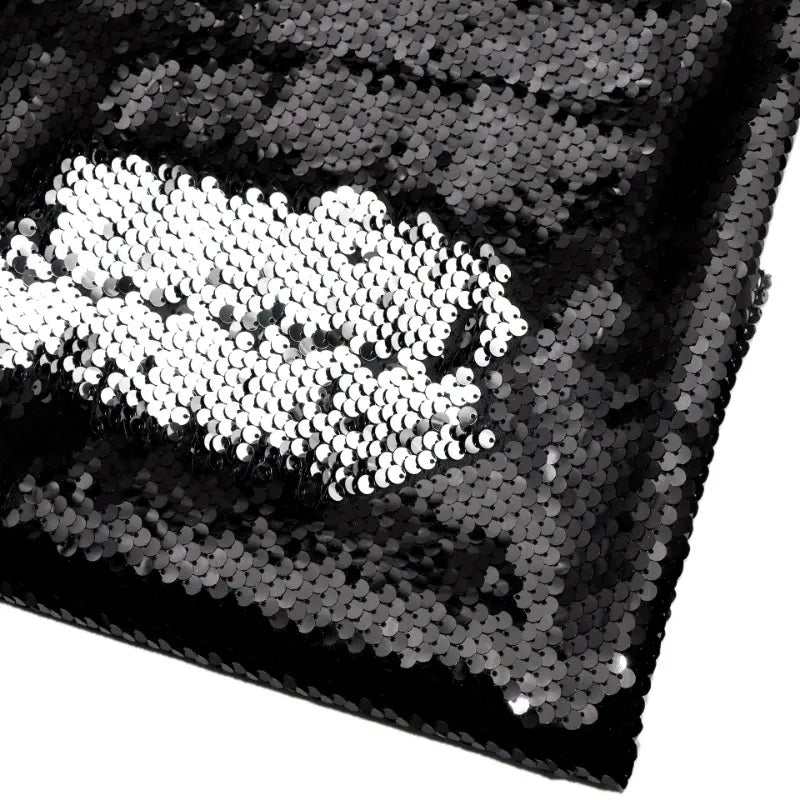 Un morceau plié du tissu pailleté bicolore réversible au mètre de Longan Craft France montre principalement des paillettes noires, avec une pièce argentée brossée créant un contraste audacieux sur le fond sombre.