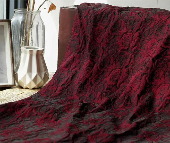 Un Longan Craft France Tissu brocart jacquard rose au mètre, présentant un motif de roses rouges et noires, est drapé sur une surface à côté d'un vase en verre, d'un vase géométrique blanc et d'un tableau encadré.