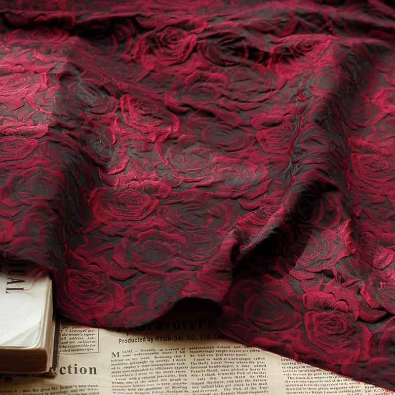 Gros plan sur le Tissu brocart jacquard rose au mètre de Longan Craft France, un jacquard ombré rouge et noir avec un motif de rose en relief, drapé sur une surface où l'on peut voir une partie d'un livre fermé et d'un journal sous le bord.