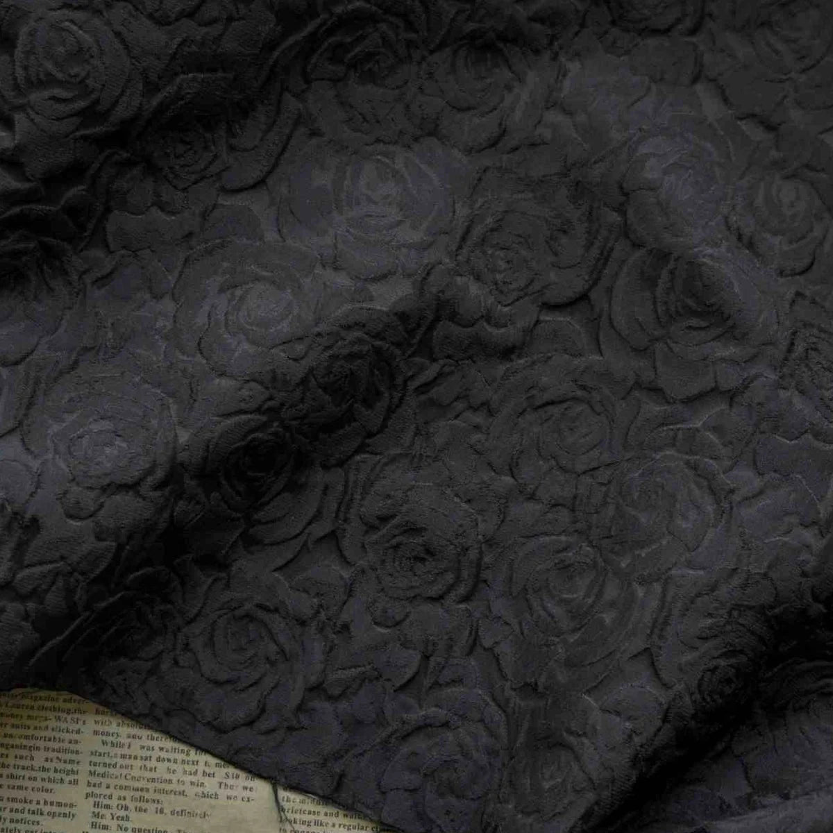 Un gros plan du Tissu brocart jacquard rose au mètre de Longan Craft France montre son motif floral rouge et noir gaufré, avec des roses texturées sur le tissu et un aperçu du journal en dessous dans le coin inférieur gauche.