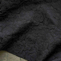 Un gros plan du Tissu brocart jacquard rose au mètre de Longan Craft France montre son motif floral rouge et noir gaufré, avec des roses texturées sur le tissu et un aperçu du journal en dessous dans le coin inférieur gauche.