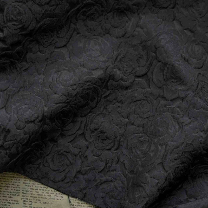 Un gros plan du Tissu brocart jacquard rose au mètre de Longan Craft France montre son motif floral rouge et noir gaufré, avec des roses texturées sur le tissu et un aperçu du journal en dessous dans le coin inférieur gauche.