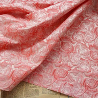 Tissu brocart jacquard rose au mètre de Longan Craft France, avec motif floral en relief façon roses, exposé sur des pages de livre ou journal. Les détails du tissu dégradé rouge et noir sont bien visibles.