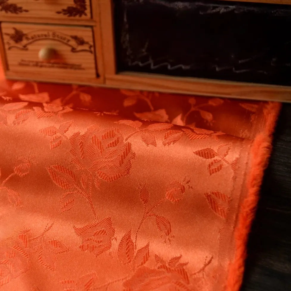 Gros plan sur le tissu satin jacquard rose de Longan Craft France, qui présente un motif floral et se drape devant un tiroir en bois muni de boutons en laiton. Ce textile est idéal pour la couture.
