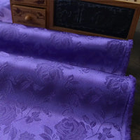 Gros plan sur le Tissu satin jacquard rose au mètre de Longan Craft France, un tissu de satin à motifs floraux idéal pour la couture, présenté devant une petite commode en bois et un tableau noir.