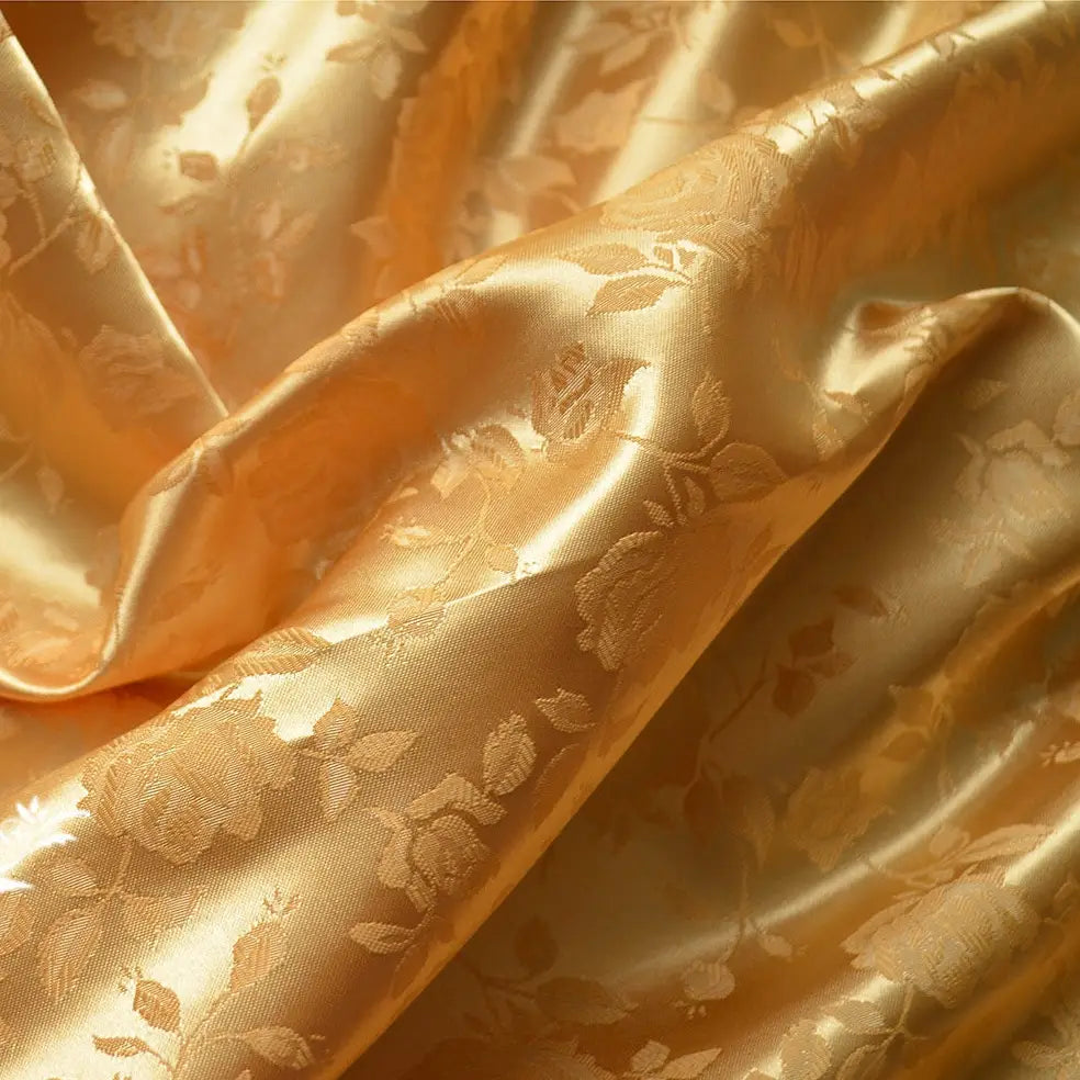 Un gros plan du Tissu satin jacquard rose au mètre de Longan Craft France montre sa finition dorée et brillante et son motif floral tissé. Le textile légèrement froissé crée des ombres élégantes, idéales pour la couture.