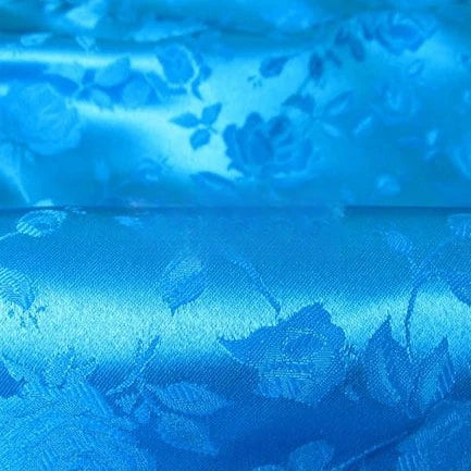 Gros plan sur le Tissu satin jacquard rose au mètre de Longan Craft France : un tissu de satin bleu brillant avec des motifs floraux gaufrés - roses et feuilles - offrant une surface texturée et réfléchissante, idéale pour la confection de robes.