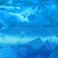 Gros plan sur le Tissu satin jacquard rose au mètre de Longan Craft France : un tissu de satin bleu brillant avec des motifs floraux gaufrés - roses et feuilles - offrant une surface texturée et réfléchissante, idéale pour la confection de robes.