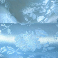 Un gros plan du Tissu satin jacquard rose au mètre de Longan Craft France montre un tissu de satin bleu clair brillant avec des motifs floraux roses tissés, réfléchissant sous une lumière douce - idéal pour la couture et les créations élégantes.
