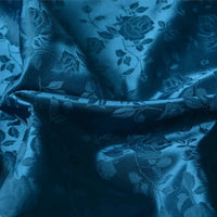 Gros plan sur le "Tissu satin jacquard rose au mètre" de Longan Craft France, un tissu de satin bleu sarcelle avec des motifs floraux en relief, une texture lisse et légèrement brillante, et des plis souples - idéal pour la couture.