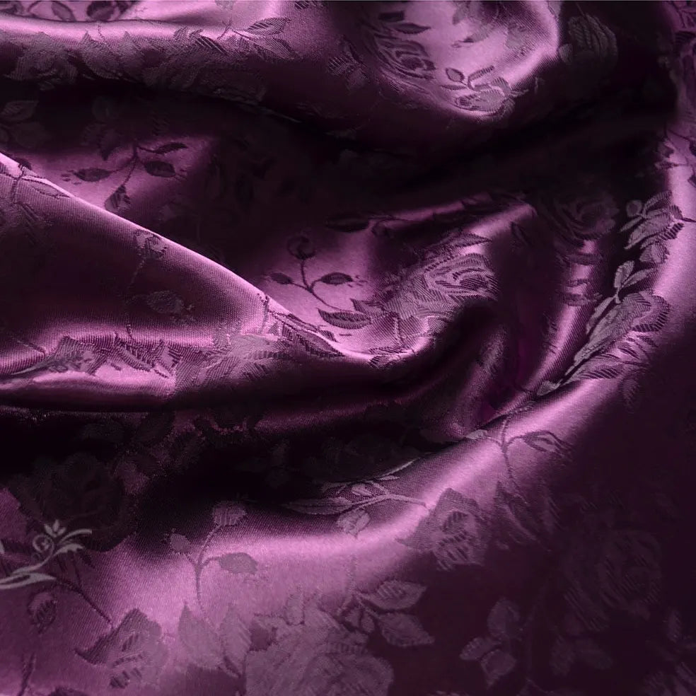 Un gros plan du Tissu satin jacquard rose au mètre de Longan Craft France montre sa surface lisse et brillante et ses subtils motifs floraux. De légers plis accentuent l'aspect satiné du tissu, idéal pour la confection de vêtements élégants.