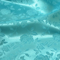 Tissu satin jacquard rose au mètre de Longan Craft France, à motifs floraux roses et feuilles, présenté dans une douce teinte bleu clair avec un aspect lisse et brillant, idéal pour la confection de robes.