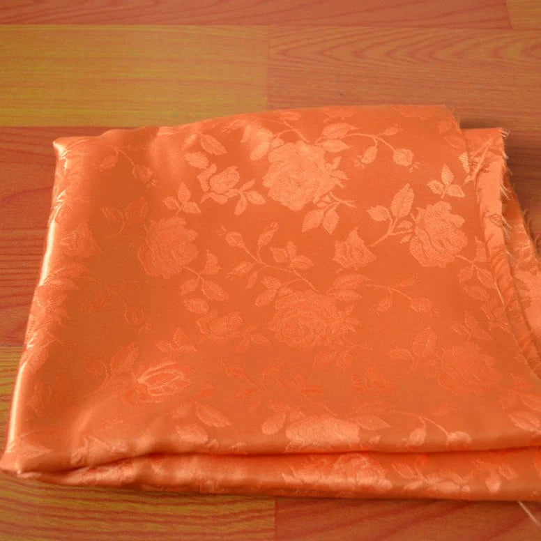 Un morceau de Tissu satin jacquard rose à motifs floraux de Longan Craft France est plié et placé sur une surface en bois rougeâtre, parfait comme textile satiné au mètre pour confection de robes.