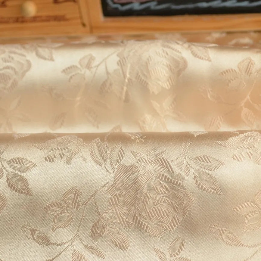 Gros plan sur le tissu satin jacquard rose au mètre de Longan Craft France, un satin beige à motifs floraux parfait pour la couture, drapé avec élégance sur un fond de bois flou.