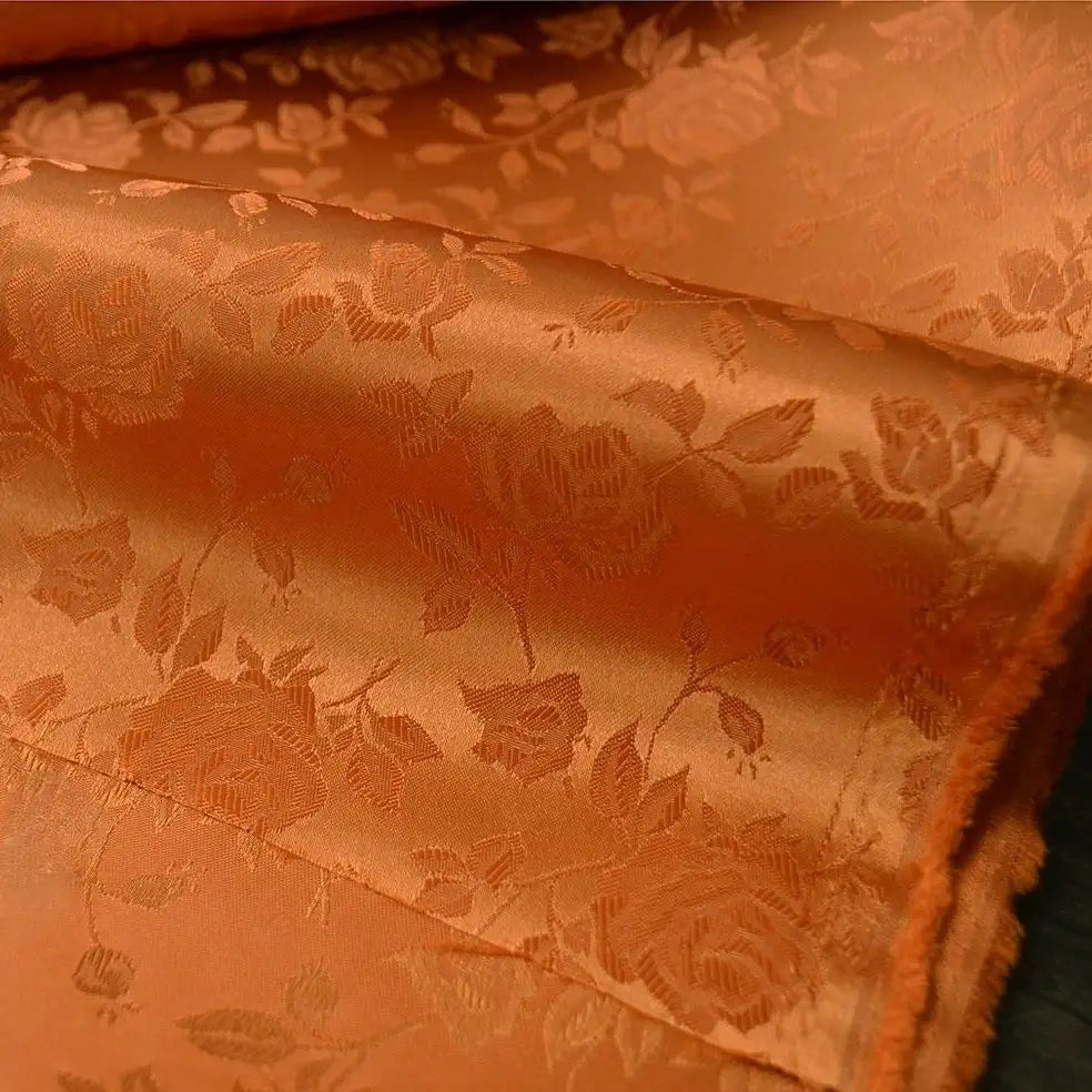 Gros plan sur le tissu satin jacquard rose au mètre de Longan Craft France, un textile de satin orange à motifs floraux avec des roses et des feuilles, montrant sa brillance subtile et sa texture détaillée, idéale pour la couture.