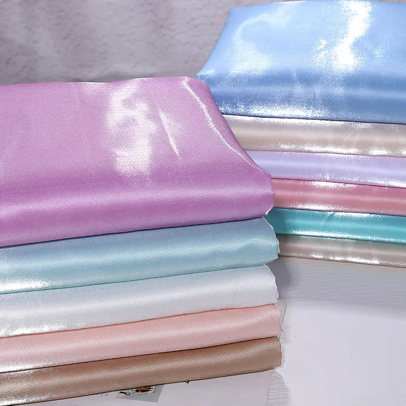 Une pile de tissus pliés en satin liquide brillant pastel - lavande, bleu, blanc, rose et beige - de Longan Craft France repose sur une surface. Idéal pour robes de demoiselle d'honneur ou tenues de cérémonie.