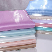 Une pile de tissus pliés en satin liquide brillant pastel - lavande, bleu, blanc, rose et beige - de Longan Craft France repose sur une surface. Idéal pour robes de demoiselle d'honneur ou tenues de cérémonie.