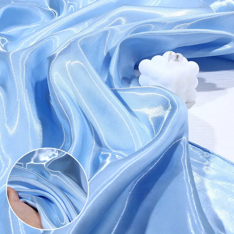Le Satin liquide brillant au mètre de Longan Craft France, tissu idéal pour robes de demoiselle d'honneur et tenues de cérémonie, est drapé sur une surface blanche avec une bougie posée dessus. Gros plan sur la texture satinée.