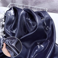 Gros plan sur le Satin liquide brillant au mètre de Longan Craft France, un tissu lustré violet foncé destiné aux robes de demoiselle d'honneur, drapé sur un panier. Un médaillon montre sa finition lisse lorsqu'on le tient dans la main.