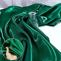 Satin liquide brillant au mètre vert émeraude de Longan Craft France drapé sur un flacon en verre transparent ; un médaillon montre une main pinçant ce tissu satin pour robes de demoiselle d'honneur pour mettre en valeur sa texture luxueuse et sa brillance.