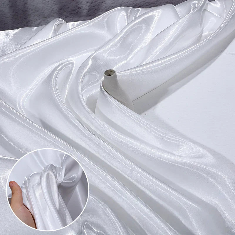 Un rouleau de Satin liquide brillant au mètre de Longan Craft France est déroulé sur une surface plane ; un médaillon montre une main qui soulève le tissu blanc lisse et brillant, idéal pour confectionner une robe de demoiselle d'honneur ou tenue de cérémonie.