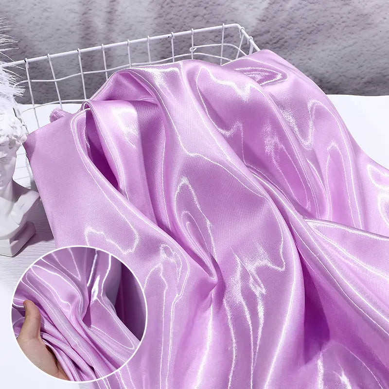 Un morceau de Satin liquide brillant au mètre de Longan Craft France, en violet clair, se drape sur un panier en fil de fer blanc, sa texture brillante et lisse reflétant la lumière. Un médaillon montre une main tenant cet élégant tissu satin pour robes de demoiselle d'honneur.