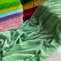 Une pile de Longan Craft France Tissu velours de soie au mètre de couleurs assorties est posée à côté d'un grand morceau de velours vert clair, mettant en valeur la texture et la brillance luxueuses idéales pour les peignoirs, la couture, l'artisanat et la tapisserie.