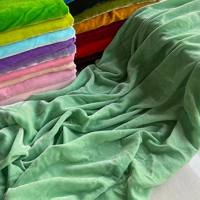 Une pile de Longan Craft France Tissu velours de soie au mètre de couleurs assorties est posée à côté d'un grand morceau de velours vert clair, mettant en valeur la texture et la brillance luxueuses idéales pour les peignoirs, la couture, l'artisanat et la tapisserie.