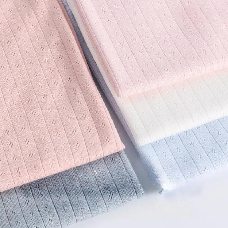 Quatre pièces de tissu jersey pointelle coton simple face de Longan Craft France, idéal pour confectionner des bodys et des pyjamas pour bébé, sont présentées dans des coloris rose pastel, blanc, bleu clair et gris avec de petits motifs d'œillets.