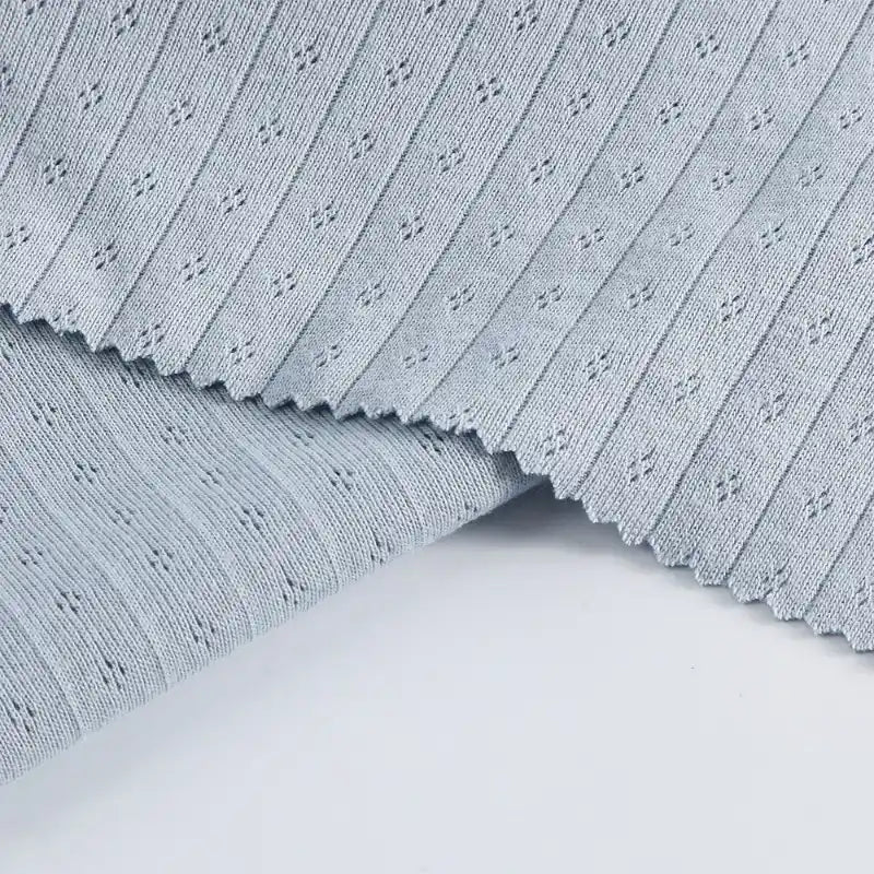 Le tissu jersey pointelle coton simple face au mètre gris clair de Longan Craft France présente une texture finement côtelée et des motifs floraux en pointillés. Il est présenté en couches avec un bord rosé, idéal pour les projets de body et de pyjama pour bébé.