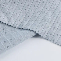 Le tissu jersey pointelle coton simple face au mètre gris clair de Longan Craft France présente une texture finement côtelée et des motifs floraux en pointillés. Il est présenté en couches avec un bord rosé, idéal pour les projets de body et de pyjama pour bébé.