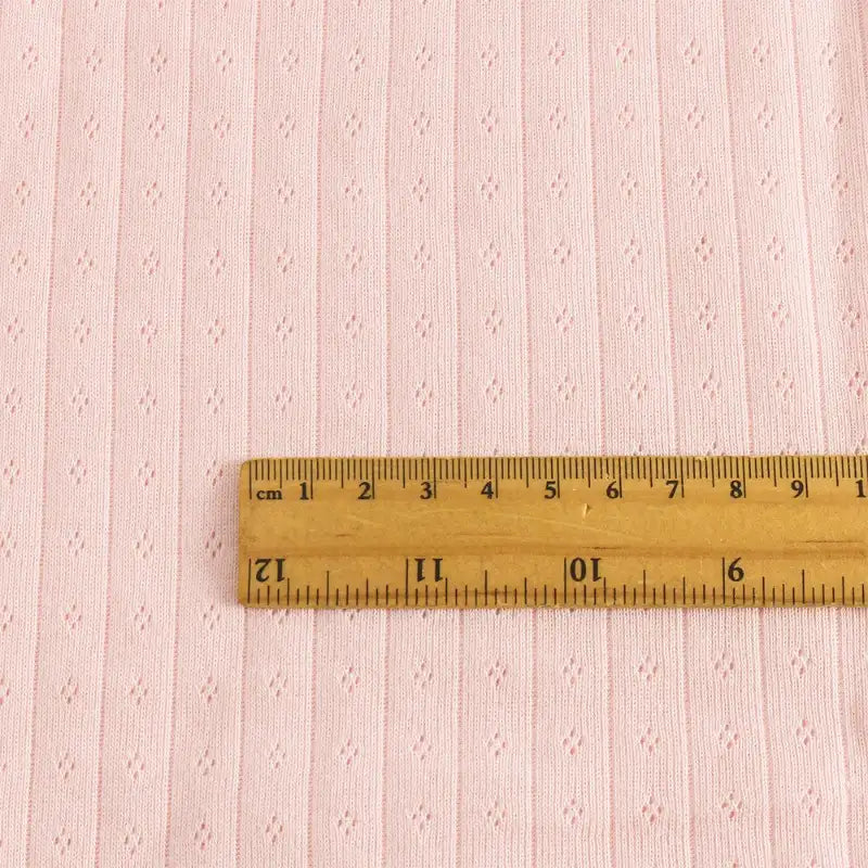Une règle en bois en pouces et centimètres repose sur le Tissu jersey pointelle coton simple face au mètre de Longan Craft France, un tissu rose pâle subtilement rayé et orné de losanges, idéal pour confectionner des bodys et des pyjamas pour bébé.