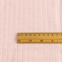 Une règle en bois en pouces et centimètres repose sur le Tissu jersey pointelle coton simple face au mètre de Longan Craft France, un tissu rose pâle subtilement rayé et orné de losanges, idéal pour confectionner des bodys et des pyjamas pour bébé.