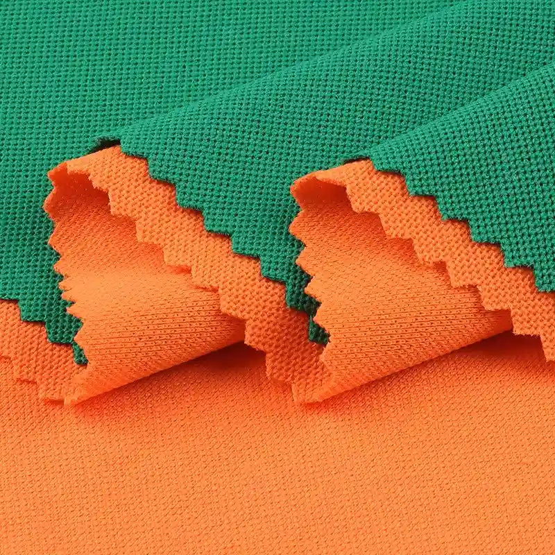 Deux pièces de Longan Craft France Tissu piqué motif œil d'oiseau à fil simple, vert et orange, sont superposées avec des bords en zigzag. Le tissu respirant est plié pour mettre en valeur les deux couleurs et sa texture œil d'oiseau, idéale pour les polos et les t-shirts d'été DIY.