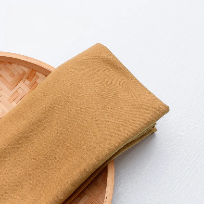 Un morceau du tissu en jersey simple au mètre, un jersey polyester-coton multicolore pour vêtements décontractés, de Longan Craft France, est plié sur un plateau rond en bambou tressé sur une surface blanche.