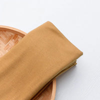 Un morceau du tissu en jersey simple au mètre, un jersey polyester-coton multicolore pour vêtements décontractés, de Longan Craft France, est plié sur un plateau rond en bambou tressé sur une surface blanche.
