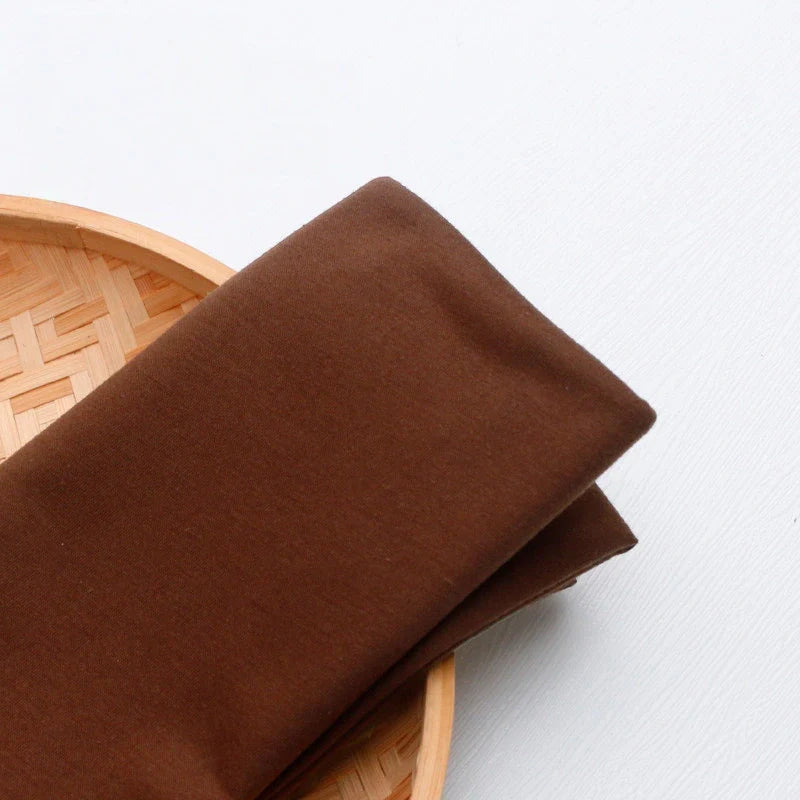 Un morceau de tissu multicolore en jersey de polyester-coton de Longan Craft France est plié sur un plateau rond en bambou tressé, posé sur une surface blanche.