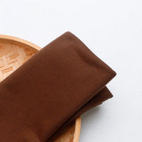 Un morceau de tissu multicolore en jersey de polyester-coton de Longan Craft France est plié sur un plateau rond en bambou tressé, posé sur une surface blanche.