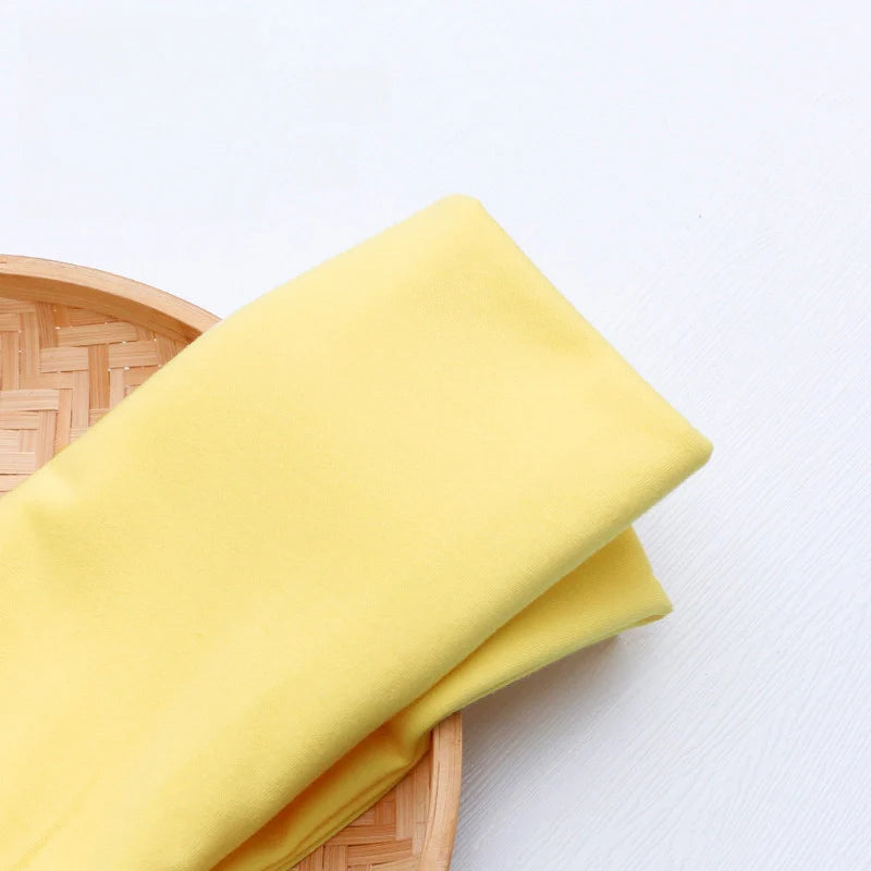 Un morceau de Tissu en jersey simple au mètre jaune clair de Longan Craft France repose sur le bord d'un plateau rond en bambou sur une surface blanche.