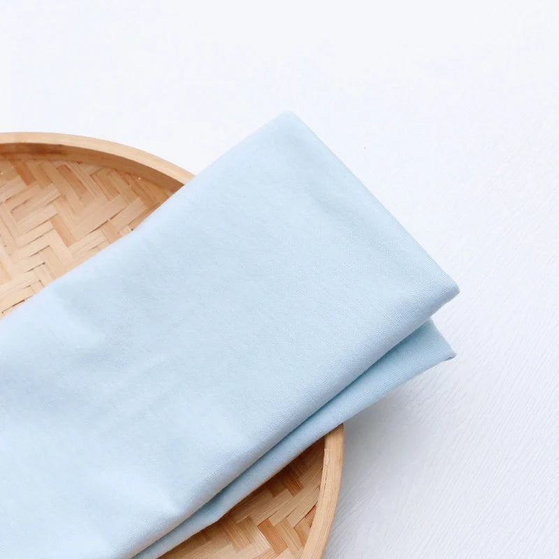 Un "Tissu en jersey simple au mètre" bleu clair de Longan Craft France est plié proprement dans un panier rond tressé sur une surface blanche.