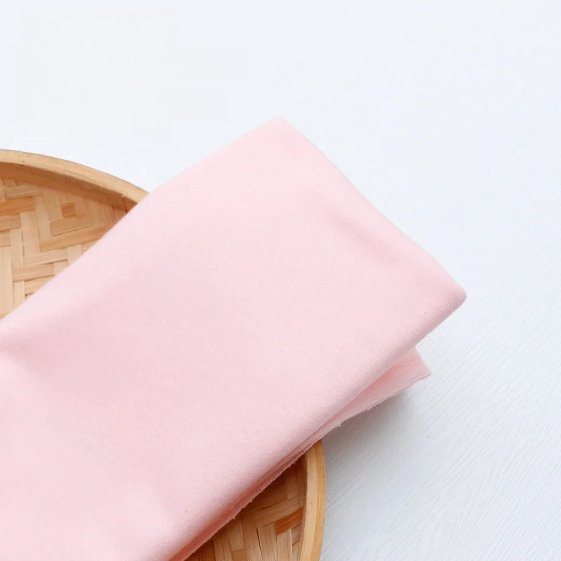 Un morceau de Tissu en jersey simple au mètre de Longan Craft France, en polyester-coton rose clair, est plié sur le bord d'un plateau rond en bambou posé sur une surface blanche.