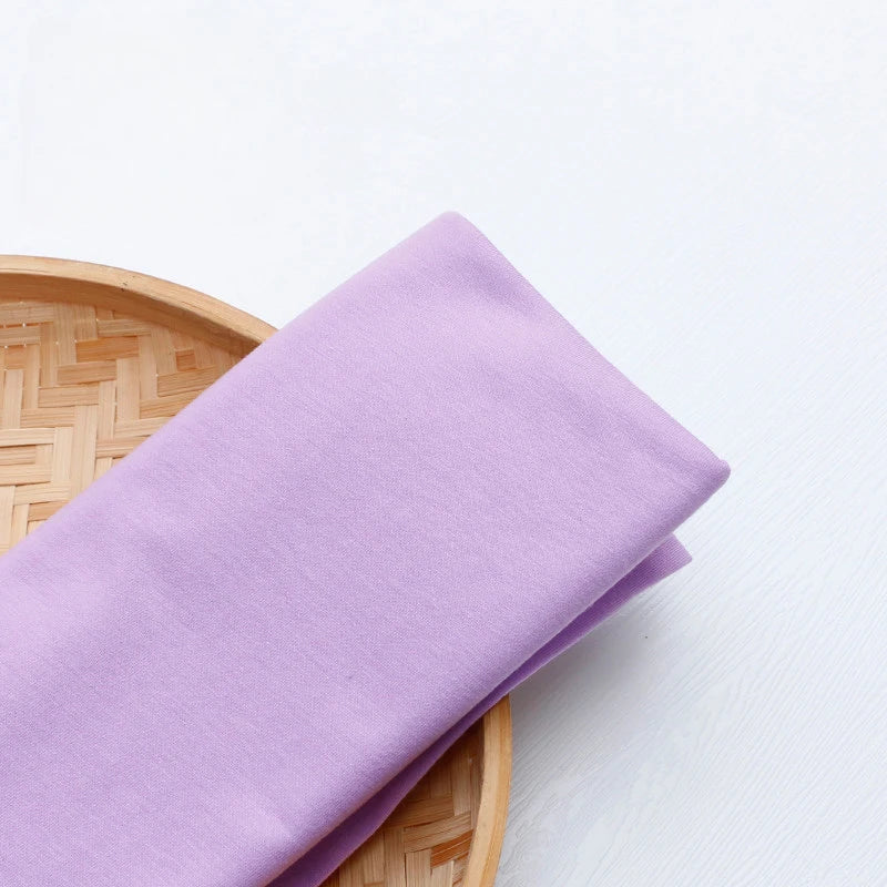 Un morceau de tissu en jersey simple au mètre de Longan Craft France, un tissu multicolore en polyester-coton pour une tenue décontractée, est plié et placé sur un plateau rond en bambou tressé sur une surface blanche.