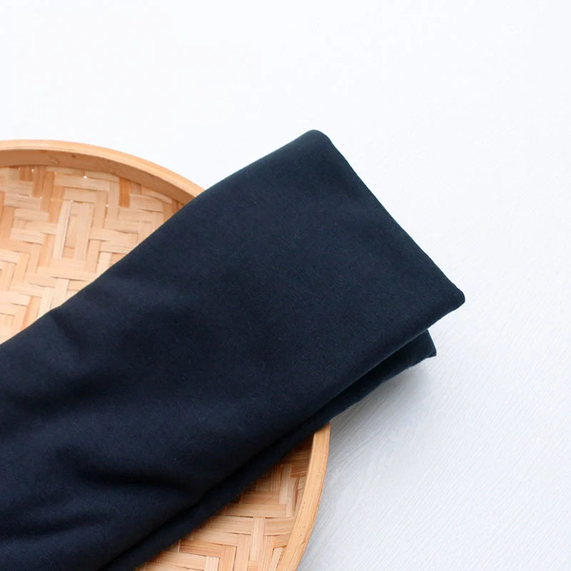 Un morceau de "Tissu en jersey simple au mètre" de Longan Craft France, un tissu multicolore en jersey de polyester et de coton pour les tenues décontractées, est présenté plié sur un plateau rond en bambou tressé sur une surface blanche.