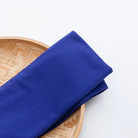 Un morceau plié de Tissu en jersey simple au mètre de Longan Craft France en bleu royal repose sur un plateau rond en bambou, posé sur une surface blanche.