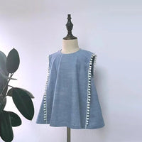 Le Patron gratuit de blouse sans manches de Longan Craft France est un haut bleu sans manches avec des pompons blancs sur les côtés, présenté sur une robe avec une plante à feuilles en arrière-plan. Un patron de couture PDF ludique et accessible aux débutantes pour l'été.