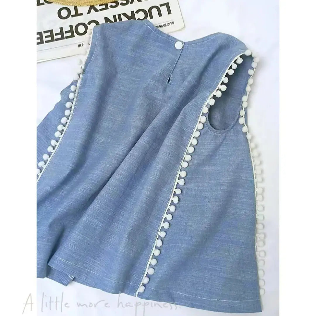 Une robe bleue sans manches avec des pompons blancs, présentée à plat sur une surface blanche avec un fond de journal - découvrez le Patron gratuit de blouse sans manches PDF de Longan Craft France, idéal pour votre prochain projet de couture d'été.