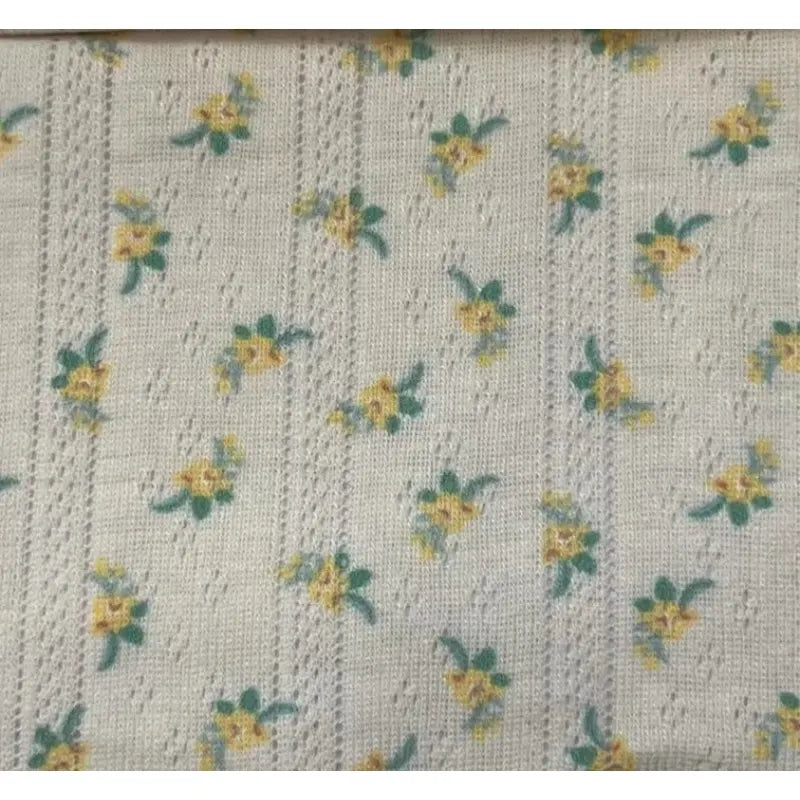 Gros plan sur le tissu pointelle à petites fleurs au mètre de Longan Craft France, un tricot imprimé d'été avec des rangées de petits motifs floraux jaunes et verts et des détails verticaux en forme de dentelle, idéal pour les robes et les gilets d'enfants.