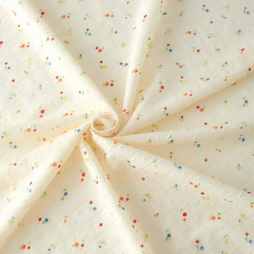 Le Tissu plumetis à petites fleurs au mètre de Longan Craft France est un tissu de coton crème au motif subtilement texturé et aux petits imprimés floraux multicolores, parfait pour des vêtements confortables.